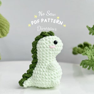 May include: A green crocheted dinosaur toy with a light green body and dark green spikes. The toy is sitting on a white surface. The text "No Sew PDF PATTERN Dinosaur" is written above the toy.