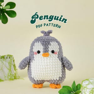 May include: A handmade crochet penguin with a gray body, white belly, and orange feet and beak. The penguin has black eyes and pink cheeks. The text "Penguin PDF Pattern" is written in green cursive above the penguin.