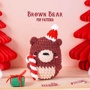 May include: A crocheted brown bear wearing a red and white Santa hat, holding a candy cane. The bear has a light beige belly and a black nose. The image includes the text "BROWN BEAR PDF PATTERN". Red paper Christmas trees and a small green gift box are also visible.