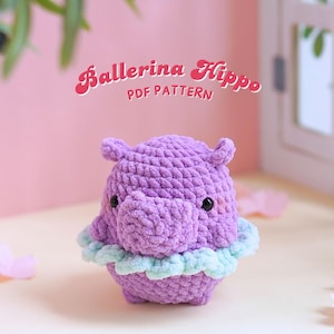 Puede incluir: Un hipopótamo bailarina de crochet morado con un tutú azul claro y verde. El hipopótamo está sentado sobre una superficie blanca con pétalos de flores rosas esparcidos a su alrededor. El texto "Ballerina Hippo PDF PATTERN" está escrito en letras rosas y blancas sobre el hipopótamo.