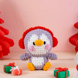 May include: A handmade crochet penguin doll with a gray body, white chest, and orange feet and beak. The penguin wears red earmuffs and has pink cheeks. Small gift boxes with red bows are in the foreground. The background is a light pink.