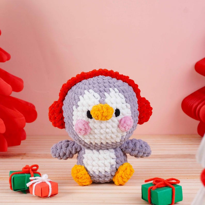 May include: A handmade crochet penguin doll with a gray body, white chest, and orange feet and beak. The penguin wears red earmuffs and has pink cheeks. Small gift boxes with red bows are in the foreground. The background is a light pink.