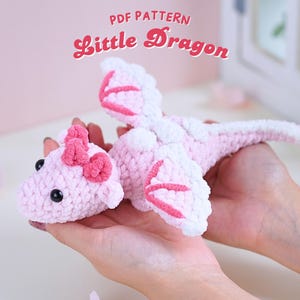 Könnte beinhalten: Ein rosa gehäkelter Drache mit weißen Akzenten und einer rosa Schleife. Der Drache hat einen langen Schwanz und Flügel. Der Text "PDF PATTERN Little Dragon" befindet sich über dem Drachen.