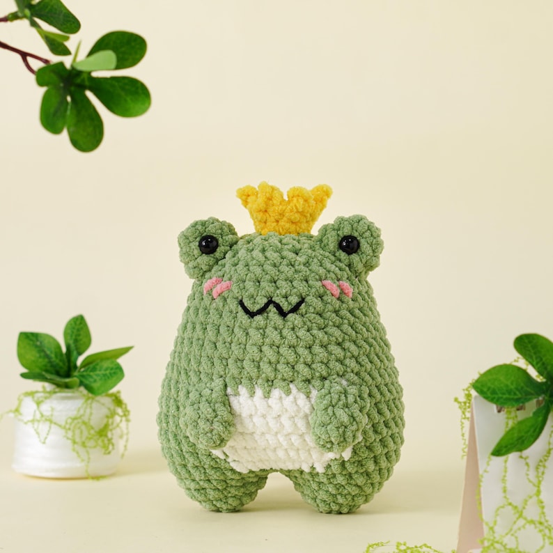 May include: A handmade, green crochet frog with a yellow crown. The frog has black eyes, pink cheeks, and a white belly. The plush toy is a whimsical, soft sculpture, perfect for a child's room or as a decorative item.