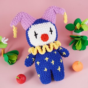 May include: A handmade blue and white clown doll with a red nose, black eyes, and yellow stars. The doll has a yellow ruffled collar and purple hat with yellow accents. The doll is surrounded by decorative fruit and greenery on a pink background.