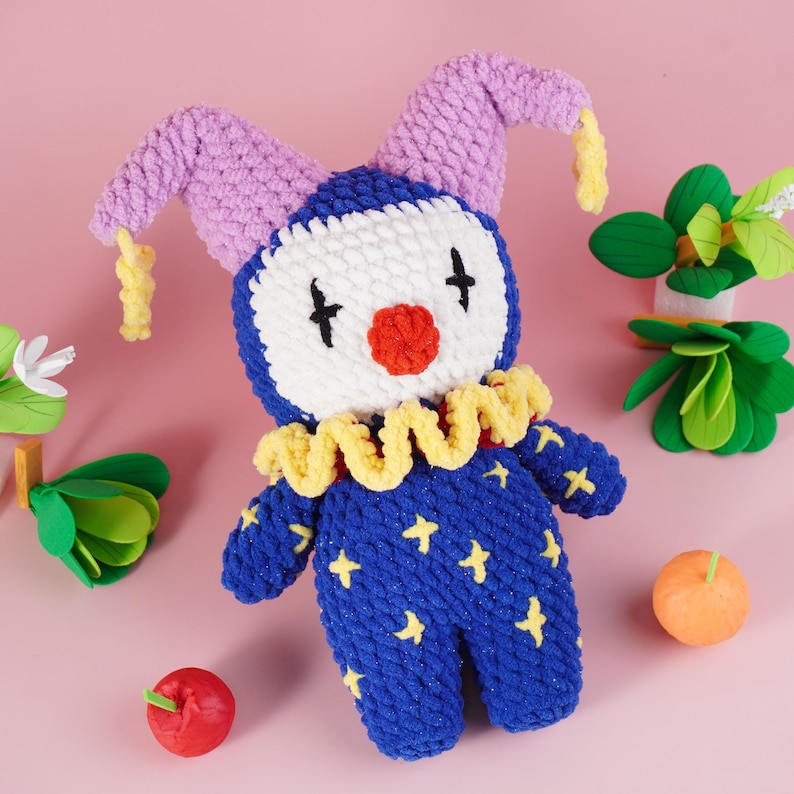 May include: A handmade blue and white clown doll with a red nose, black eyes, and yellow stars. The doll has a yellow ruffled collar and purple hat with yellow accents. The doll is surrounded by decorative fruit and greenery on a pink background.