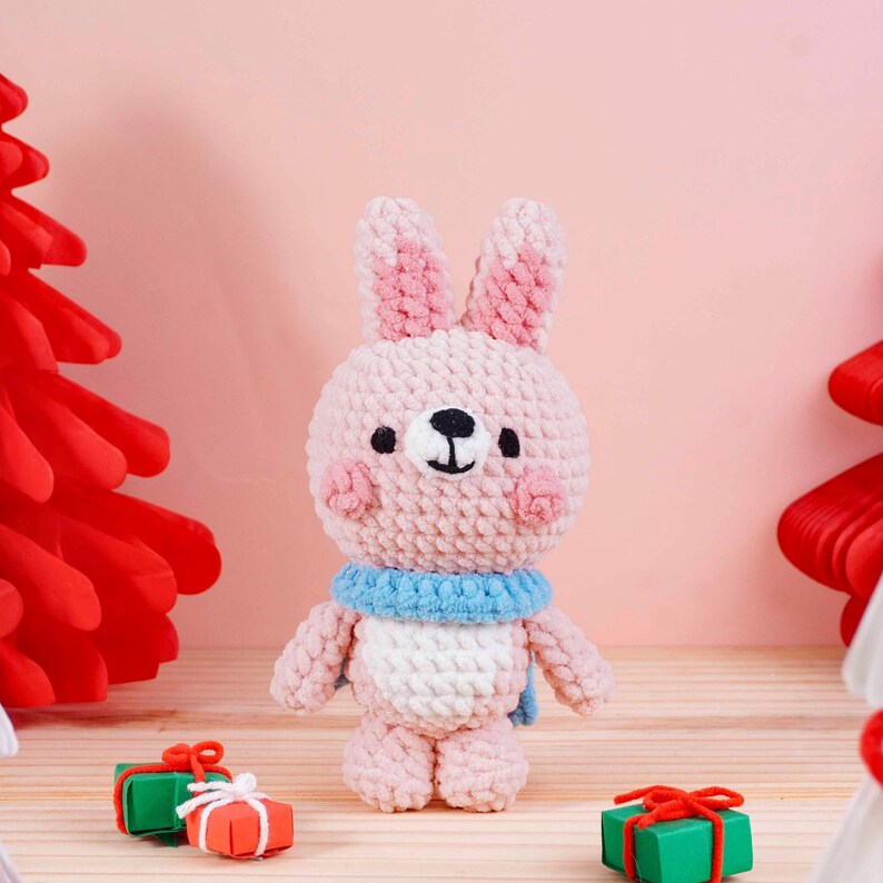 May include: A handmade pink crochet bunny with a blue scarf and white chest. The bunny has pink inner ears and cheeks, a black nose and eyes. Two small gift boxes are in the foreground. The background is a light pink.
