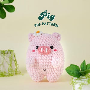 Puede incluir: Un juguete de amigurumi de cerdo de crochet rosa con una flor blanca en la cabeza. El juguete está sentado sobre una superficie de color amarillo claro. El texto "Pig PDF Pattern" está escrito en letras verdes sobre el juguete.