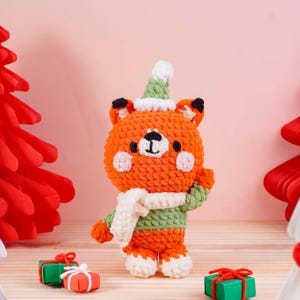 May include: A handmade orange fox plushie wearing a green and white hat, scarf, and sweater. The fox has black eyes and ears, and pink cheeks. Two small wrapped gifts are in the foreground. The background is a light pink color.