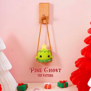 Puede incluir: Adorno de fantasma de pino verde tejido a ganchillo con una estrella amarilla, colgado de una clavija de madera. El texto "Pine Ghost PDF Pattern" está debajo. El fondo incluye árboles de Navidad de papel rojo y blanco y pequeñas cajas de regalo.