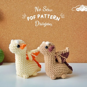 May include: Two crocheted dragon toys, one white with orange wings and one brown with brown wings. The text "No Sew PDF PATTERN Dragon" is on a corkboard background.