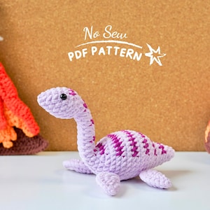 Könnte beinhalten: Ein gehäkelter lila Dinosaurier mit rosa Streifen. Der Dinosaurier liegt auf einer weißen Oberfläche. Der Text "No Sew PDF PATTERN" ist im Hintergrund sichtbar.