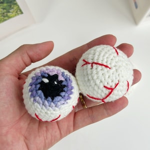 May include: Two crocheted eyeballs, one with a purple and blue iris and the other with red stitching.