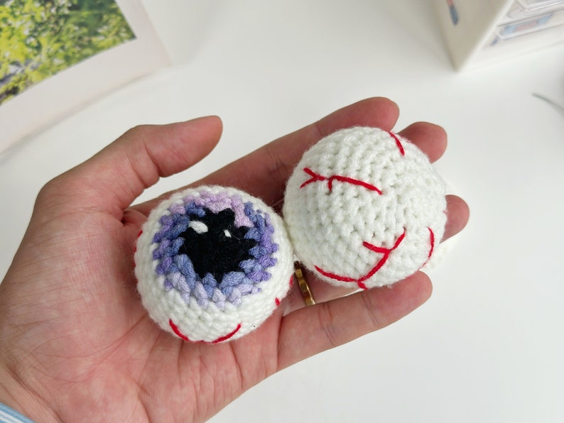 May include: Two crocheted eyeballs, one with a purple and blue iris and the other with red stitching.
