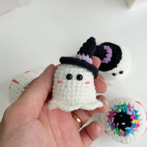 May include: Three crocheted Halloween decorations: a ghost wearing a purple and black witch hat, a white eyeball with red and blue details, and a colorful eyeball with a black pupil.