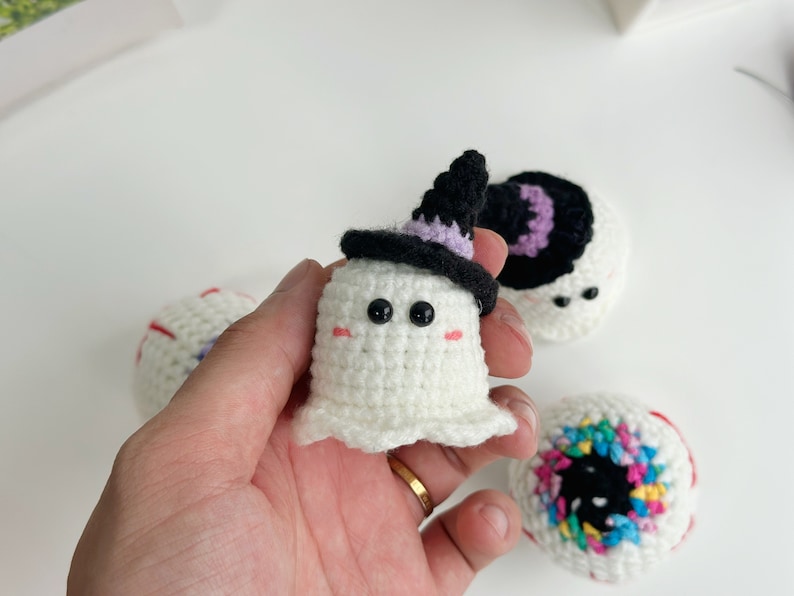 May include: Three crocheted Halloween decorations: a ghost wearing a purple and black witch hat, a white eyeball with red and blue details, and a colorful eyeball with a black pupil.