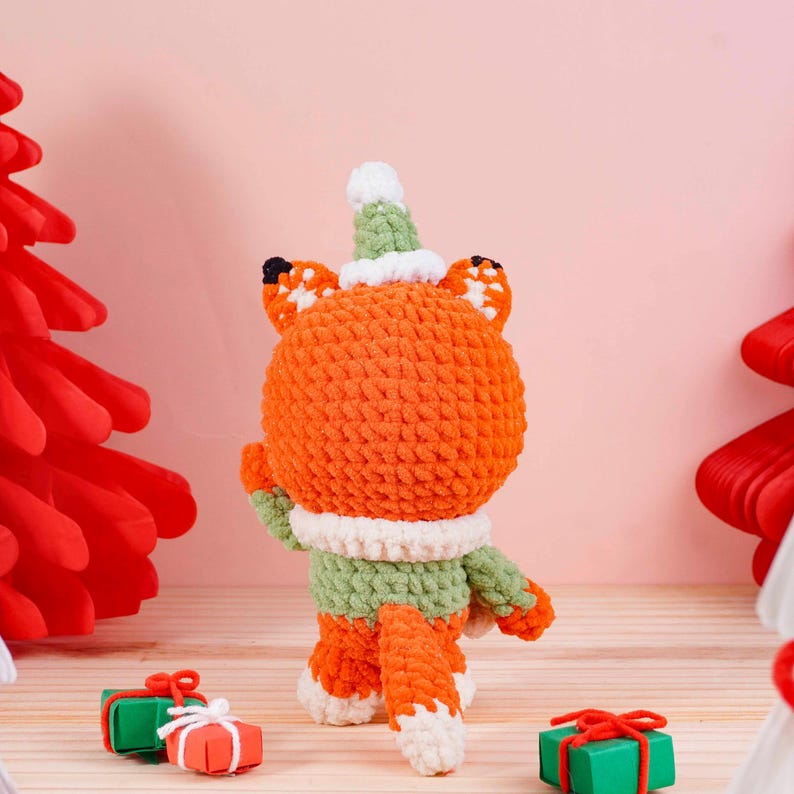 May include: A handmade orange fox plushie wearing a green and white scarf and hat. The fox has black accents on its ears and is standing near small wrapped gifts. The plushie is a festive holiday decoration.
