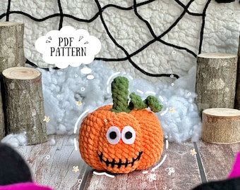 May include: A crocheted orange pumpkin with a green stem and googly eyes. The pumpkin has a stitched-on mouth and is sitting on a white fluffy surface. The text "PDF PATTERN" is in a thought bubble above the pumpkin.