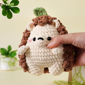 May include: A handmade crochet hedgehog toy in cream, brown, and green. The hedgehog has a cream-colored body, brown spines, and a green leaf on its head. It has black eyes and pink cheeks. The toy is soft and plush.