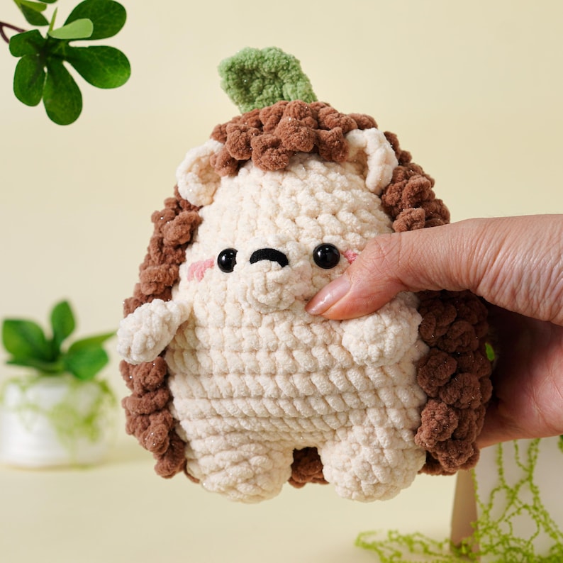 May include: A handmade crochet hedgehog toy in cream, brown, and green. The hedgehog has a cream-colored body, brown spines, and a green leaf on its head. It has black eyes and pink cheeks. The toy is soft and plush.