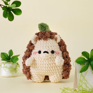 May include: A handmade crochet hedgehog plush toy. The toy features a cream-colored body, brown spiky back, and a green leaf on top. It has small arms and legs, with a cute face, including pink cheeks and black eyes. The toy is designed to be soft and cuddly.