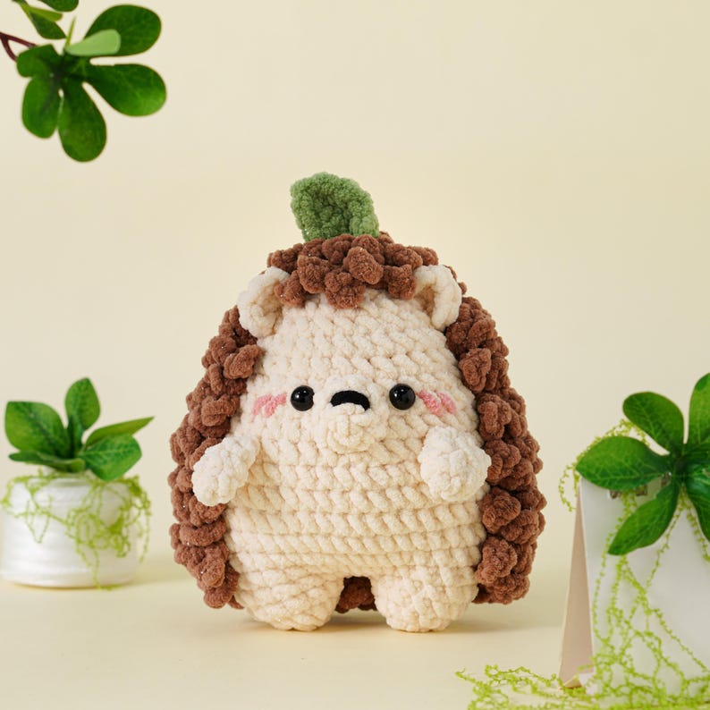 May include: A handmade crochet hedgehog plush toy. The toy features a cream-colored body, brown spiky back, and a green leaf on top. It has small arms and legs, with a cute face, including pink cheeks and black eyes. The toy is designed to be soft and cuddly.