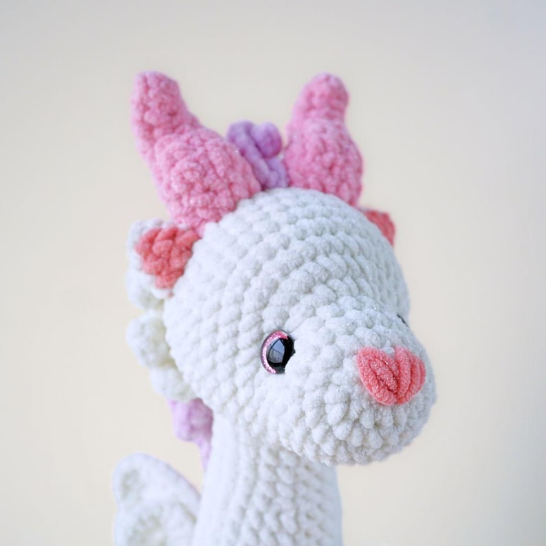 May include: A white crocheted dragon with pink horns and a pink nose. The dragon has black eyes and a sweet expression.