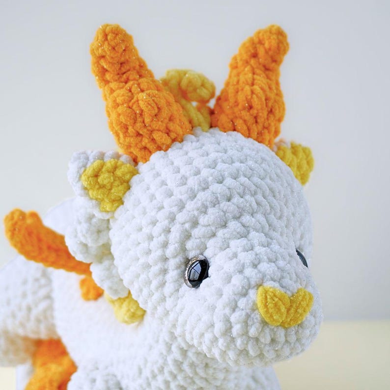 May include: White crocheted plush dragon with orange and yellow horns and accents. The dragon has black eyes and a yellow nose.