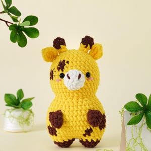 May include: A handmade yellow crochet giraffe with brown spots and accents. The stuffed animal has a white muzzle, small black eyes, and blush-colored cheeks. The giraffe is a soft, plush toy, perfect for children.