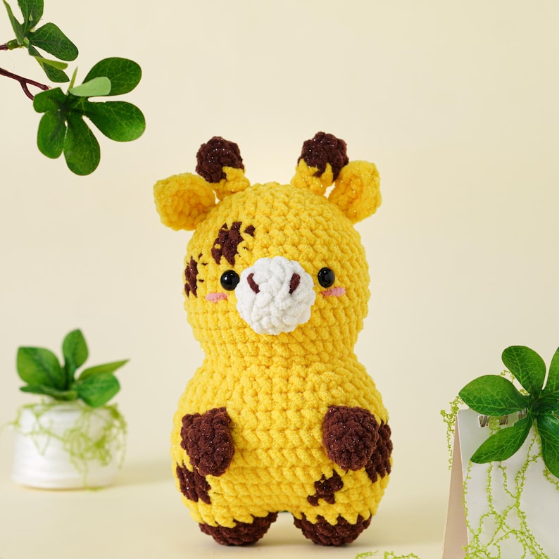 May include: A handmade yellow crochet giraffe with brown spots and accents. The stuffed animal has a white muzzle, small black eyes, and blush-colored cheeks. The giraffe is a soft, plush toy, perfect for children.