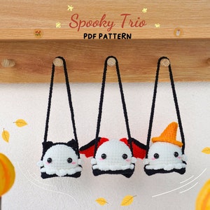 以下が含まれることがあります： 木製の壁にフックで吊るされた、かぎ針編みで作られた幽霊のぬいぐるみ3体。それぞれの幽霊は、コウモリ、悪魔、魔女の帽子という異なるハロウィーンの衣装を着ています。ぬいぐるみは白で、目は黒く、口も黒いです。背景は白い壁で、床には秋の落ち葉が散らばっています。木製の壁には「Spooky Trio PDF Pattern」という文字が書かれています。