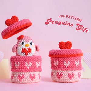 May include: PDF pattern for crocheting a penguin gift box. The box is pink and white with a heart design. The lid is red with a heart on top. The penguin is pink and white with a red heart on its chest. The text "PDF PATTERN Penguins Gift" is in the upper right corner of the image.