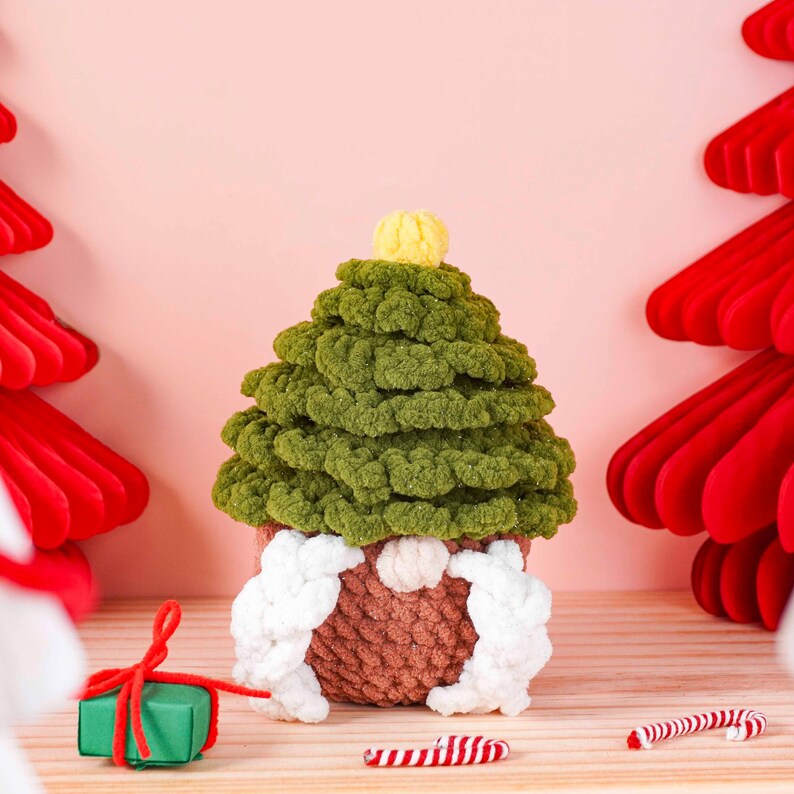May include: A handmade Christmas gnome in the shape of a green tree. The gnome has a brown body, white beard and arms, and a yellow pom-pom on top. A small green gift box with a red bow and candy canes are in the foreground.