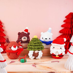 May include: A collection of handmade Christmas ornaments. The set includes a reindeer, bear, polar bear, Christmas tree, and Santa Claus, all crocheted in festive colors. The ornaments are arranged on a wooden surface with decorative paper trees.