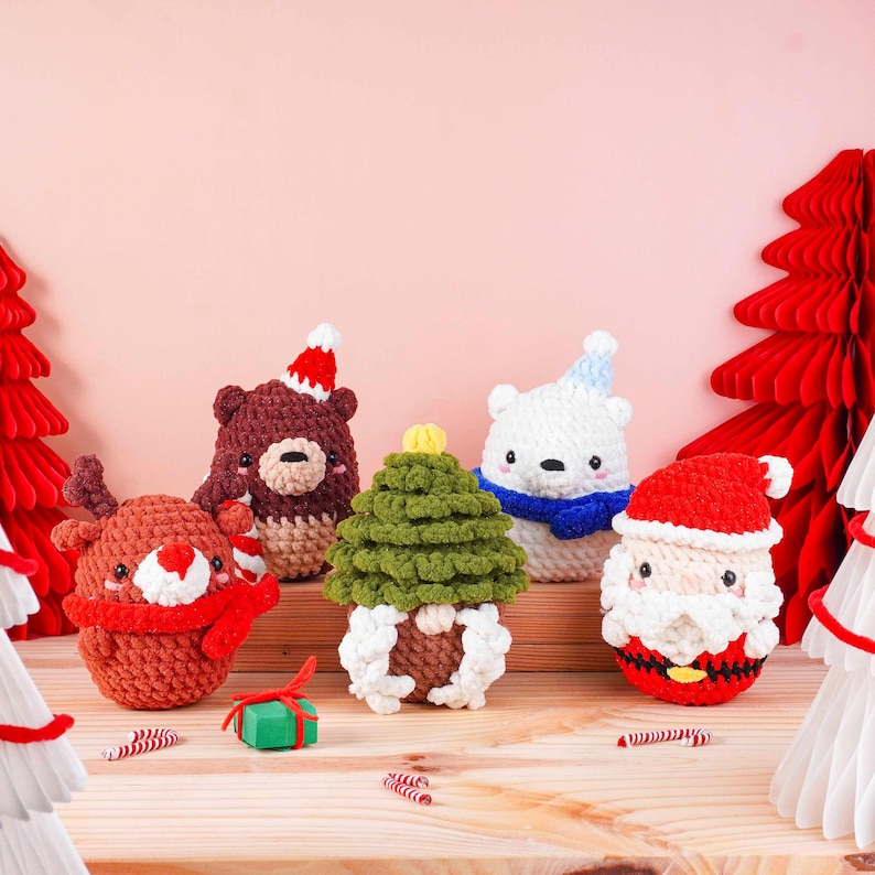 May include: A collection of handmade Christmas ornaments. The set includes a reindeer, bear, polar bear, Christmas tree, and Santa Claus, all crocheted in festive colors. The ornaments are arranged on a wooden surface with decorative paper trees.