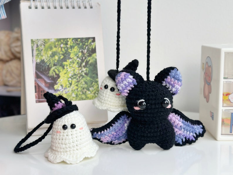 May include: Two crocheted Halloween ornaments, a white ghost wearing a black witch hat and a black bat with purple and blue wings.
