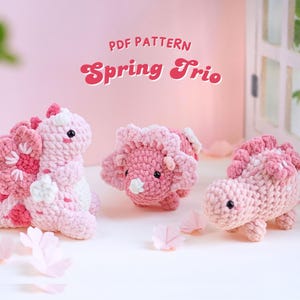 Può includere: Tre animali amigurumi all'uncinetto rosa: un dinosauro, un triceratopo e uno stegosauro. Gli animali sono tutti seduti su una superficie bianca con petali di fiori rosa sparsi intorno a loro. Il testo "PDF PATTERN Spring Trio" è nella parte superiore dell'immagine.
