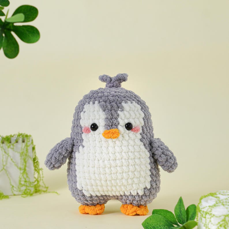 May include: A handmade crochet penguin plushie. The penguin has a gray head and wings, a white chest, and orange feet and beak. It has black eyes and pink cheeks. The plushie is standing on a light yellow surface.