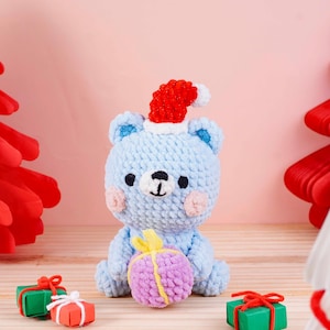 May include: A light blue crocheted bear wearing a red Santa hat, holding a purple and yellow gift. The bear has pink cheeks and black eyes. Small wrapped gifts are in the foreground. The background is a light pink.