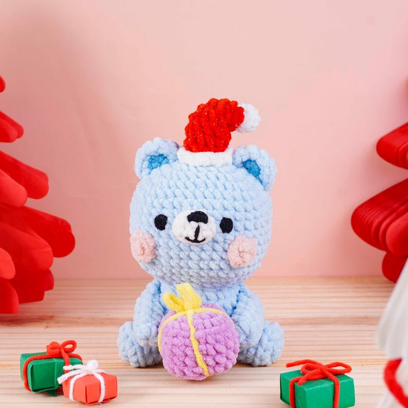 May include: A light blue crocheted bear wearing a red Santa hat, holding a purple and yellow gift. The bear has pink cheeks and black eyes. Small wrapped gifts are in the foreground. The background is a light pink.
