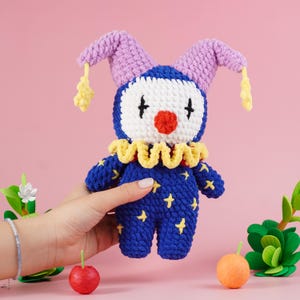 May include: A handmade blue and yellow crochet clown doll with a white face, red nose, and black cross eyes. The doll has purple ears and yellow star accents. The doll is held in a hand against a pink background.