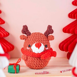 May include: A handmade, crocheted reindeer ornament with a red scarf and antlers. The reindeer has a white nose and black eyes. The ornament is sitting on a wooden surface with a small green gift box and candy canes.
