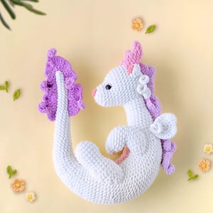 May include: A white crocheted dragon with a pink and purple tail and wings. The dragon is curled up in a sitting position.