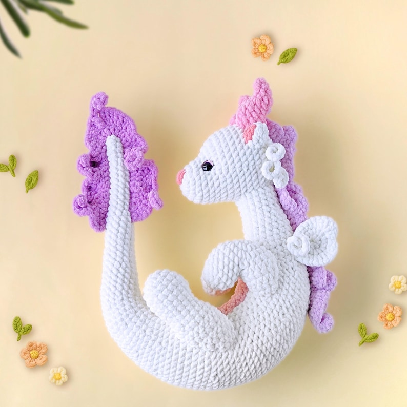 May include: A white crocheted dragon with a pink and purple tail and wings. The dragon is curled up in a sitting position.