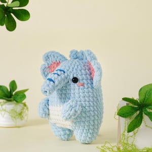 May include: A light blue crochet elephant with a white belly. The elephant has pink accents on its ears and a blue trunk. It has black eyes and a small pink mouth. The elephant is a handmade item.
