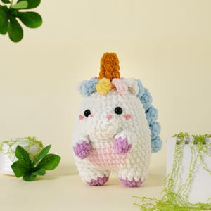 May include: A handmade, white crochet unicorn toy with a textured body. It features a golden horn, a blue, yellow, and pink mane, and purple hooves. The unicorn has black eyes and pink cheeks, creating a whimsical appearance.