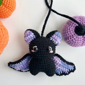May include: A crocheted black bat with purple and blue wings. The bat has pink cheeks and black eyes. The bat is hanging from a purple crocheted ball.