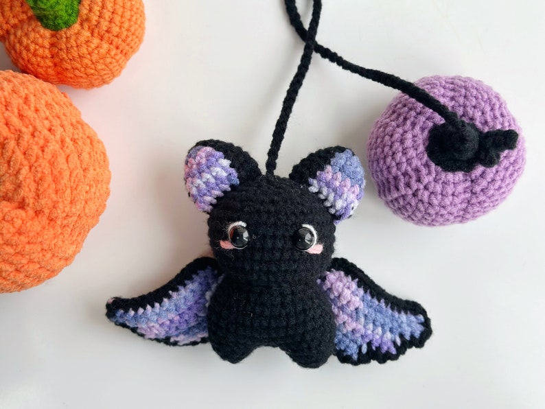 May include: A crocheted black bat with purple and blue wings. The bat has pink cheeks and black eyes. The bat is hanging from a purple crocheted ball.