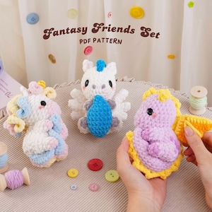 May include: Three crocheted plush toys from the Fantasy Friends Set PDF Pattern. One is white with pastel accents, one is white with blue, and one is purple with yellow wings. Small buttons and thread spools are scattered nearby.
