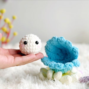 May include: A white crocheted plush toy with black eyes and a pink mouth, sitting inside a blue crocheted shell with white crocheted accents.